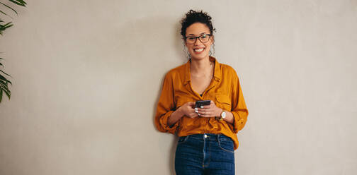 Porträt einer glücklichen jungen Frau, die an einer Wand steht. Eine Unternehmerin, die ein Mobiltelefon in der Hand hält, schaut in die Kamera und lächelt. - JLPSF08405
