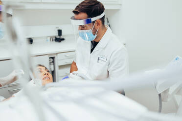 Ein Zahnarzt behandelt eine Patientin in einer Zahnklinik. Ein männlicher Arzt mit Maske und Gesichtsschutz bespricht die Behandlung mit einem Patienten in einer Zahnklinik. - JLPSF08411