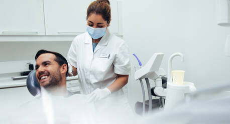 Male patient sitting in dentist's chair looking away and smiling with a female nurse putting on dental apron on the patient. Dentist assisting a patient to wear dental apron. - JLPSF08420