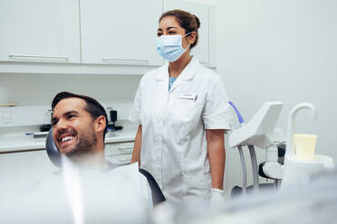 Man witting in dentist chair looking at side and smiling with a female dentist wearing face mask standing by. Dentist and patient looking at something and smiling in dental clinic. - JLPSF08421