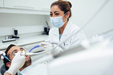 Dentist wearing face mask smiling while doing a dental treatment on male patient. Dental doctors working on a patient's teeth in clinic. - JLPSF08424