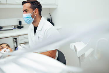 Male dentist talking with his assistant while examining female patient's teeth in clinic. Dentist in protective face mask while treating a patient in a dental office. - JLPSF08427