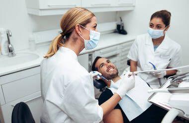 Dental doctor looking at a display monitor while treating a male patient. Female dentist wearing face mask with male patient sitting in dentist's chair and her assistant sitting by in dental clinic. - JLPSF08428