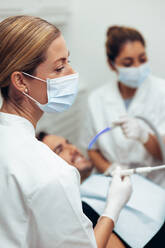 Close-up of a female dental surgeon treating a patient in clinic with her assistant in background. Dentist treating a male patient in her office. - JLPSF08430