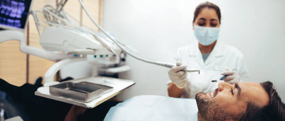 Smiling man sitting in dentist's chair receiving dental treatment from a female dentist. Male patient getting dental checkup in clinic. - JLPSF08432