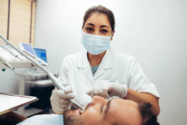 Female dentist wearing face mask looking at camera while treating a patient. Doctor doing dental treatment on man's teeth in the dentists chair. - JLPSF08435