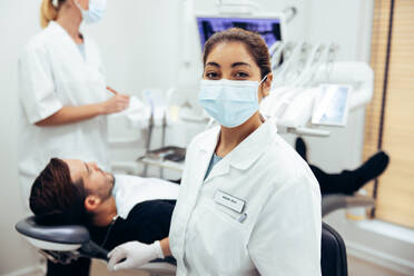 Frau bei der Arbeit in einer Zahnklinik mit einem Patienten auf einem Stuhl und einem Arzt im Hintergrund, dessen Assistentin mit Gesichtsmaske in die Kamera schaut. - JLPSF08437