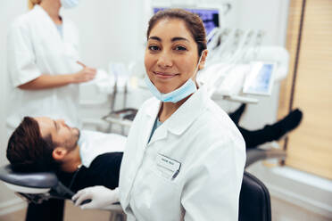 Happy female dentist looking at camera. Dental doctor in her clinic with patient in dentist's chair and nurse in background. - JLPSF08438