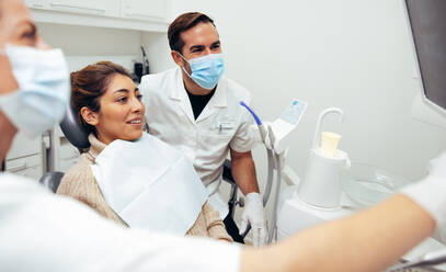 Woman sitting in dentist's chair looking at her dental x-ray with doctors around. Dentist showing x-ray to patient and explaining the procedure of treatment. - JLPSF08443