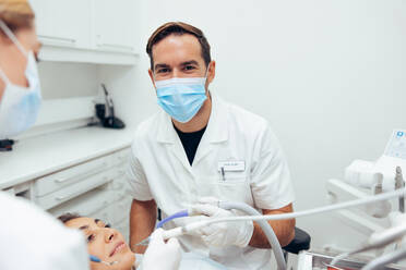 Erfolgreicher Zahnarzt bei der Arbeit an den Zähnen eines Patienten in seiner Klinik. Männlicher Zahnarzt mit Gesichtsmaske, der in die Kamera schaut, während der Patient im Stuhl sitzt und sein Assistent davor steht. - JLPSF08448