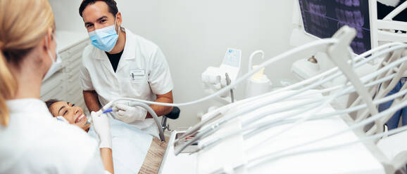 Male dentist discussing with his assistant while treating a patient in clinic. Doctors performing a dental treatment on female patient sitting in dentist's chair. - JLPSF08450