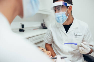 Ärzte mit Gesichtsschutz, die zwischen der zahnärztlichen Behandlung einer Patientin sprechen. Ein männlicher Zahnarzt, der einen Absaugschlauch hält, diskutiert mit seiner Assistentin während der Behandlung. - JLPSF08455