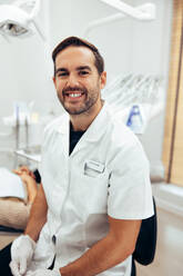 Portrait of confident dental expert in his clinic. Male dentist wearing lab coat looking at camera and smiling. - JLPSF08456