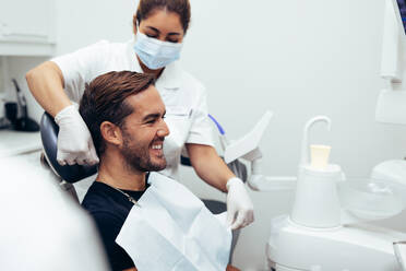 Krankenschwester nimmt einem männlichen Patienten nach einer Zahnbehandlung in einer Klinik die Schürze ab. Männlicher Patient lächelt nach seiner Zahnbehandlung. - JLPSF08472