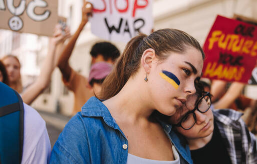 End the conflict in Ukraine. Young woman marching against war with the flag of Ukraine painted on her face. Group of multicultural young people holding an anti-war protest in the city. - JLPSF08503