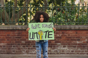 Ein schwarzes Teenager-Mädchen schaut in die Kamera und hält ein Plakat, das zur Einheit aufruft. Ein Teenager-Aktivist protestiert gegen Rassenungleichheit und Diskriminierung. - JLPSF08519