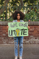 Ein junges schwarzes Mädchen blickt in die Kamera und hält ein Plakat, das zur Einheit aufruft. Eine jugendliche Aktivistin protestiert gegen Rassenungleichheit und Diskriminierung. - JLPSF08520