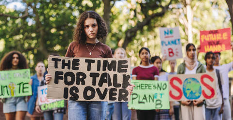 Teenager-Mädchen schaut in die Kamera, während sie einen Marsch gegen den Klimawandel anführt. Gruppe multiethnischer Jugendaktivisten, die gegen die globale Erwärmung protestieren. Junge Menschen, die sich dem globalen Klimastreik anschließen. - JLPSF08522