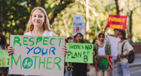 Fröhliches Teenager-Mädchen, das in die Kamera schaut, während es einen Marsch gegen die globale Erwärmung anführt. Gruppe multiethnischer Jugendaktivisten, die gegen den Klimawandel protestieren. Teenager, die sich dem globalen Klimastreik anschließen. - JLPSF08523