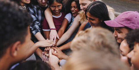 Multicultural teenagers smiling happily while putting their hands together in a huddle. Group of diverse young people symbolizing unity and teamwork. - JLPSF08567