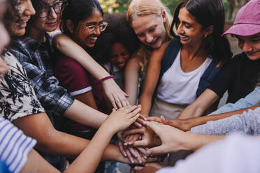 Diverse teenagers smiling cheerfully while putting their hands together in a huddle. Group of generation z youngsters symbolizing team spirit and togetherness. - JLPSF08570