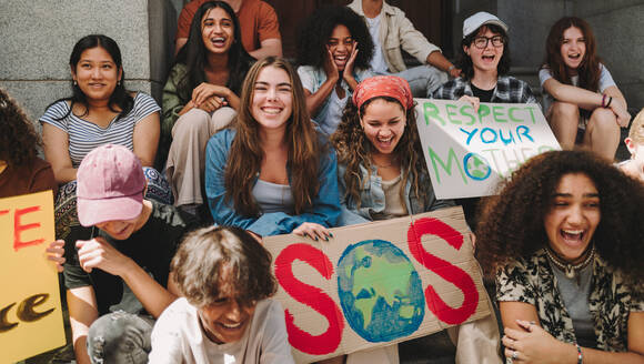 Youth environmental campaign. Group of multicultural climate activists smiling happily while sitting with posters and placards outside a building. Young people protesting against global warming. - JLPSF08591