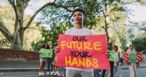 Junger Aktivist, der während einer Kundgebung zum Klimawandel ein Transparent in der Hand hält und wegschaut. Ein Jugendlicher protestiert gegen die globale Erwärmung mit einer Gruppe von Demonstranten im Hintergrund. - JLPSF08611