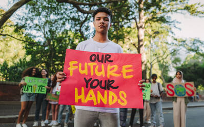 Ein Jugendlicher schaut in die Kamera, während er bei einer Kundgebung zum Klimawandel ein Transparent hält. Aktivist der Generation Z protestiert gegen die globale Erwärmung mit einer Gruppe von Demonstranten im Hintergrund. - JLPSF08613