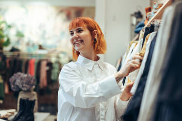 Lächelnde Einkäuferin in einem Bekleidungsgeschäft. Frau betrachtet Kleidung, die auf einem Gestell in einem Modegeschäft hängt. - JLPSF08621