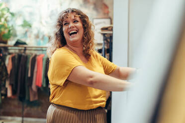 Eine Kundin lächelt, während sie Kleidung auf einem Regal in einem Geschäft betrachtet. Eine Frau kauft in einem Bekleidungsgeschäft ein. - JLPSF08627