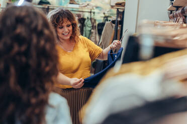 Schöne Frau hält einen Kleiderbügel mit einem neuen Kleid in einem Modegeschäft, während die Verkäuferin davor steht. - JLPSF08629