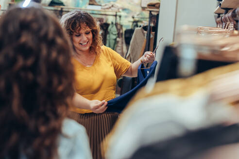 Beautiful woman holding a hanger with a new dress in the fashion store while saleswoman standing in front. Female shopper shopping in clothing store. - JLPSF08629
