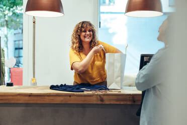 Lächelnde Besitzerin eines Bekleidungsgeschäfts, die Kleidung in eine Papiertüte packt und eine Kundin anschaut und lächelt. Weibliche Verkäuferin in einem Bekleidungsgeschäft, die hinter dem Tresen steht und Kleidung für eine Kundin in eine Papiertüte packt. - JLPSF08637