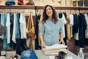 Frau ordnet Kleidung auf einem Tisch im Geschäft. Verkäuferin schaut weg und lächelt, während sie in einem Bekleidungsgeschäft arbeitet. - JLPSF08646