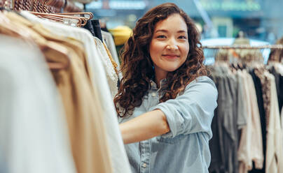 Happy young woman standing by clothing rack and smiling in shop. Female customer shopping in a clothing store. - JLPSF08652