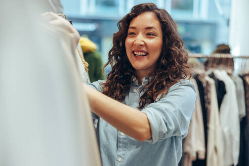 Smiling woman looking at dresses hanging in rack in shop. Woman shopping in a boutique. - JLPSF08653