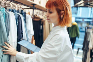 Woman with digital tablet looking at clothes hanging in store. Businesswoman working in clothing store taking stock of clothes. - JLPSF08656