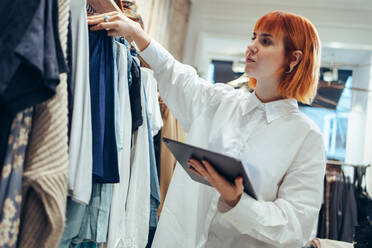 Woman manager in a fashion store holding a digital tablet and checking the shop stock. Female owner looking at clothes hanging on rack in store. - JLPSF08658