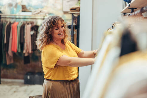 Glückliche reife Frau, die in einem Bekleidungsgeschäft arbeitet. Verkäuferin ordnet Kleidung im Regal in ihrem Modegeschäft. - JLPSF08669