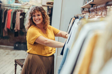 Frau, die in einem Bekleidungsgeschäft arbeitet. Weibliche Inhaberin, die wegschaut und lächelt, während sie einer Kundin in einem Modegeschäft beim Kauf hilft. - JLPSF08670