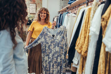 Frau hält ein Kleid und zeigt es einer Kundin in ihrem Geschäft. Besitzerin eines Bekleidungsgeschäfts hilft einer Kundin im Laden. - JLPSF08675