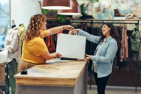 Friendly seller of clothing store giving shopping bags to satisfied female customer. Fashion store owner handing over the shopping bag to a female customer at checkout counter. - JLPSF08706