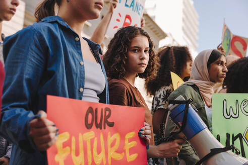 Teenage girl holding a megaphone while marching for climate justice with a group of demonstrators. Multicultural youth activists protesting against global warming and climate change. - JLPSF08819