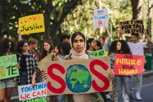 Muslim girl looking at the camera while holding a poster at a climate change rally. Group of multicultural youth activists protesting against global warming. Teenagers joining the global climate strike. - JLPSF08824