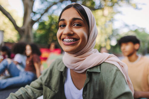 Fröhliches muslimisches Mädchen, das in die Kamera lächelt, während es mit einer Gruppe von Demonstranten bei einem Klimaprotest sitzt. Multikulturelle jugendliche Aktivisten, die sich dem globalen Klimastreik anschließen. - JLPSF08847