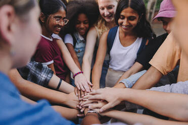 Generation Z youngsters smiling happily while putting their hands together in a huddle. Group of multicultural teenagers uiting at school event - JLPSF08853