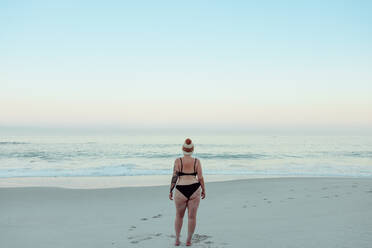 Rückansicht einer nicht erkennbaren Winterbadenden in Badekleidung am Strand. Anonyme Frau, die im Winter auf das Meerwasser schaut. Winterbadende, die allein am Meer steht. - JLPSF08865