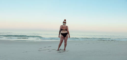 Anonyme Winterbadende in Badekleidung am Strand stehend. Rückansicht einer nicht erkennbaren Frau, die auf das Meerwasser im Winter schaut. Winterbadende, die allein am Meer steht. - JLPSF08866