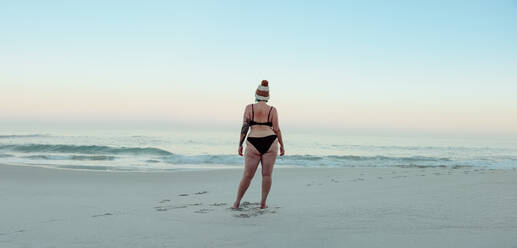 Anonymous female winter bather standing at the beach in swimwear. Rearview of an unrecognisable woman looking at the sea water in winter. Winter bather standing alone by the seaside. - JLPSF08866