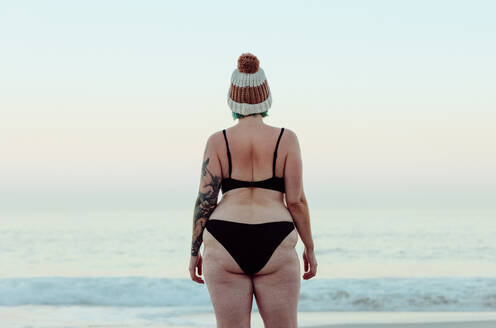 Anonyme Winterbadende in Badekleidung am Strand stehend. Rückansicht einer nicht erkennbaren Frau, die auf das Meerwasser im Winter schaut. Winterbadende, die allein am Meer steht. - JLPSF08869